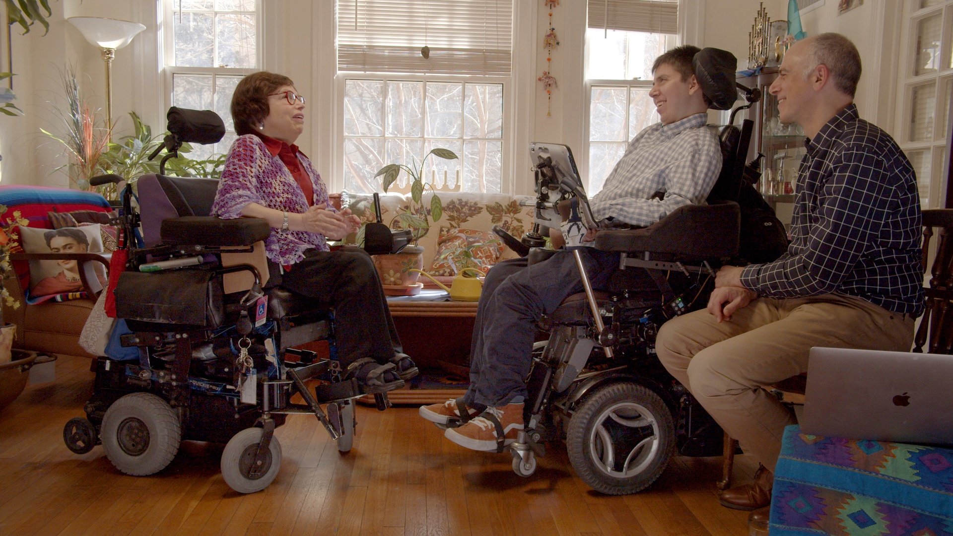 A group of three white people, one woman and two men, are sitting indoors. The woman and younger man are in motorized wheelchairs. The woman has cropped brown hair, red glasses, and is wearing a red blouse covered by a purple shawl. The younger man is wearing a checkered button down shirt, blue jeans, and sneakers. The older man is wearing a checkered button down shirt and khakis. All three individuals are smiling and appear to be in conversation.